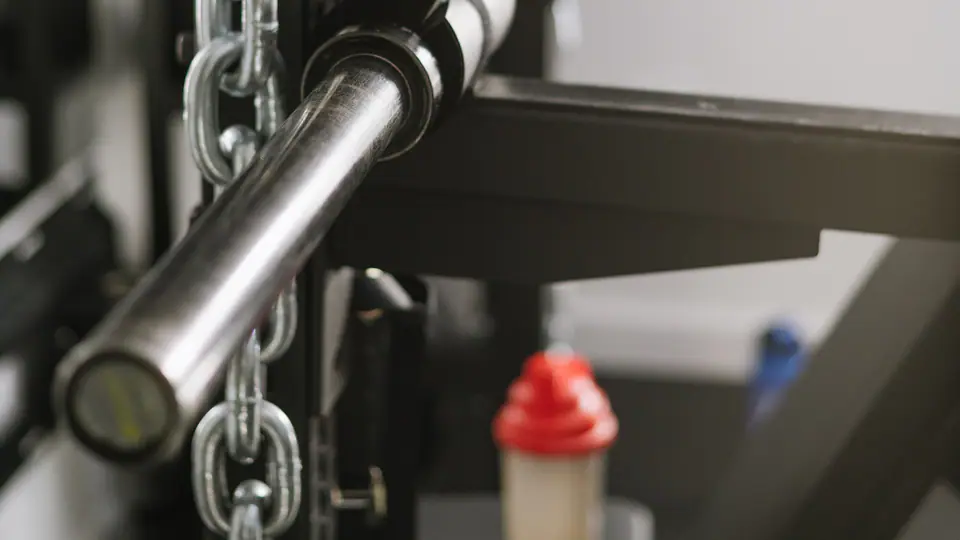 Barbell resting in a rack beside a hanging chain, with a protein shaker on the shelf below